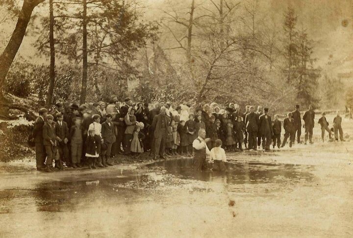 Black and white photo of a group of around fifty people stand along the riverbank, watching a baptism happening within its waters.
